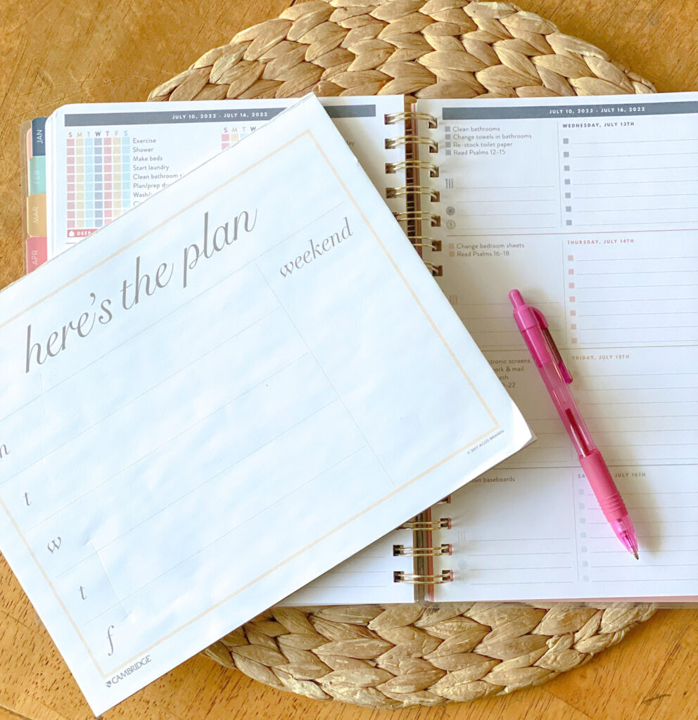Weekly planning notepad on top of a lined weekly planner and pink pen. Background straw placemat and light wooden tabletop