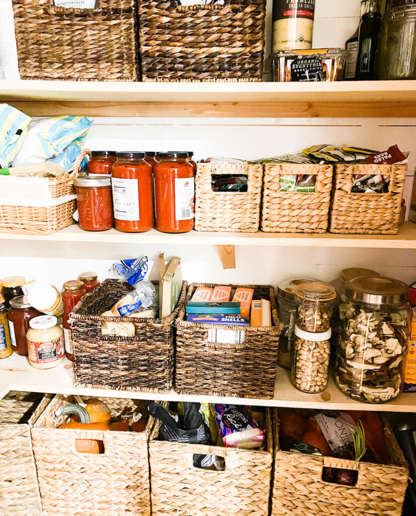 Pantry stocked with various items straw baskets for organization various glass jars filled with pantry items, pine board shelves. 