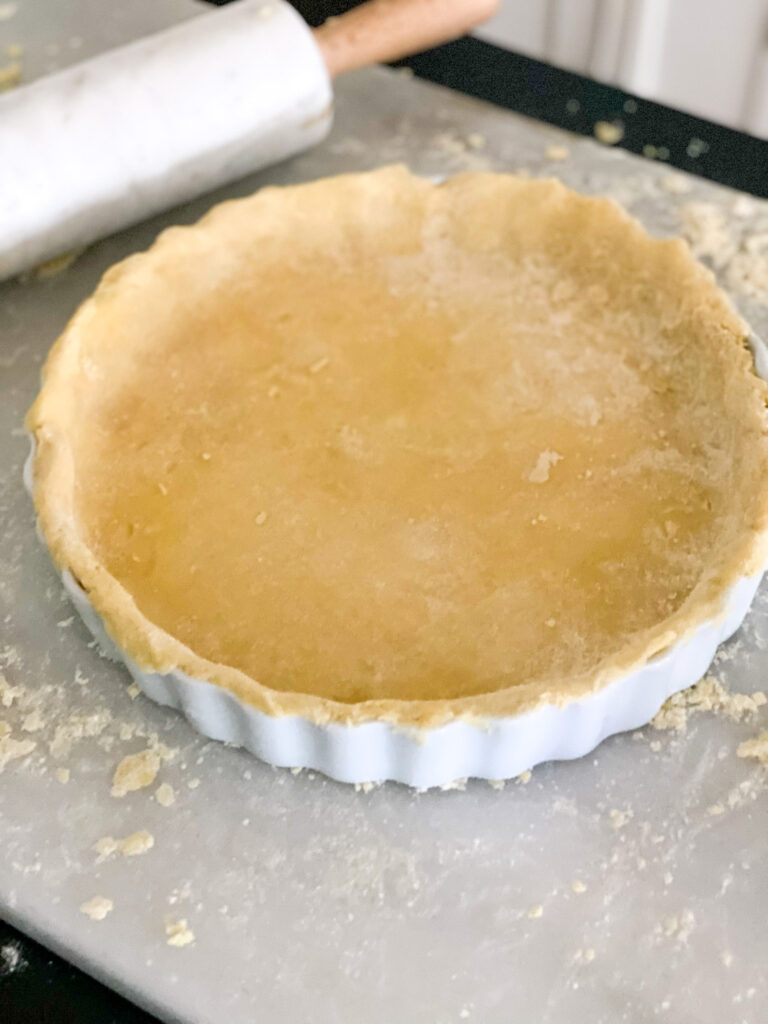 yellow colored pie dough pressed into a white pie pan surrounded my pie crust crumbs on top of a white marble pastry board white marble rolling pin with wooden handles laying toward the side