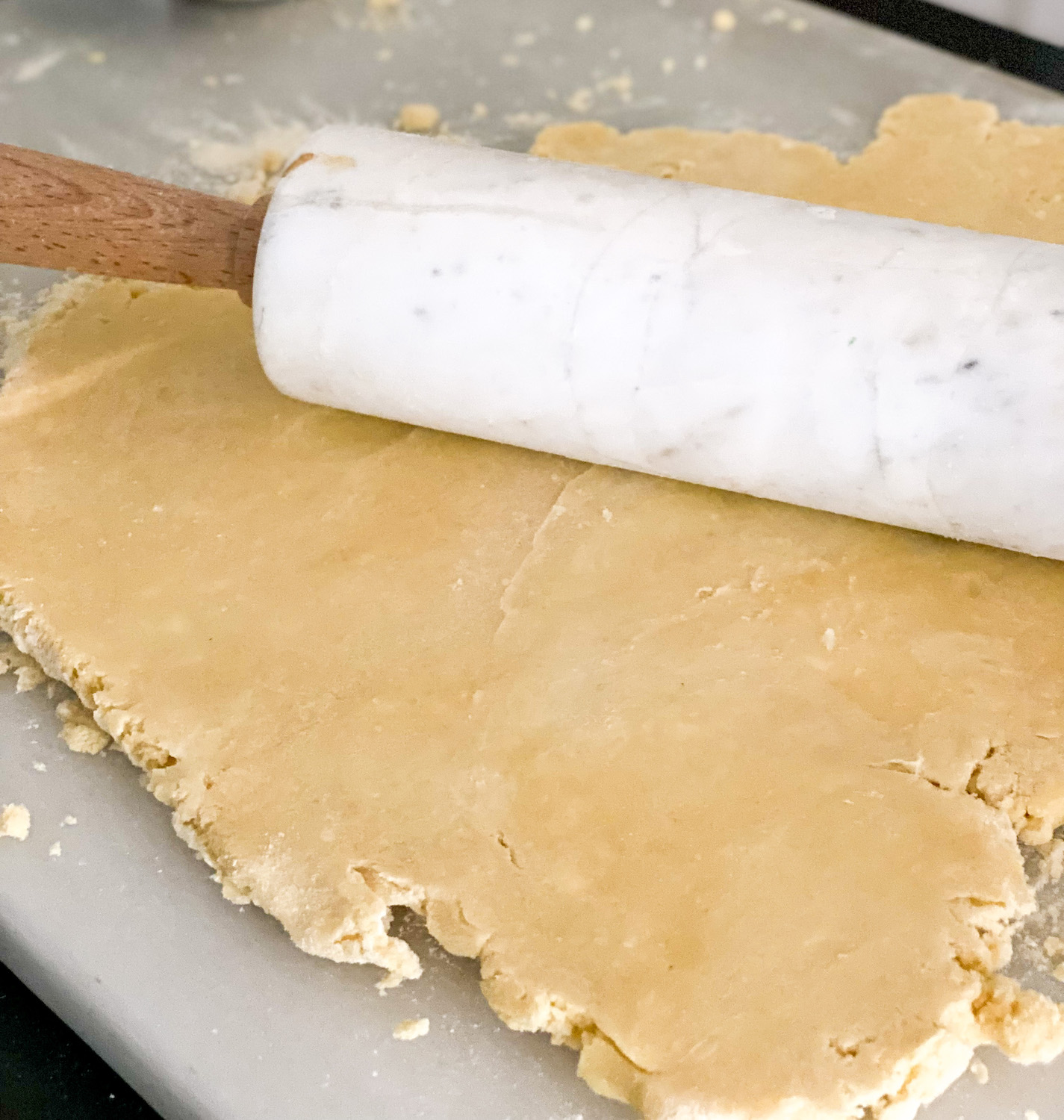 Marble rolling pin with wooden handles on top of yellow pie dough, surrounded by flour and dough crumbs. background marble dough board
