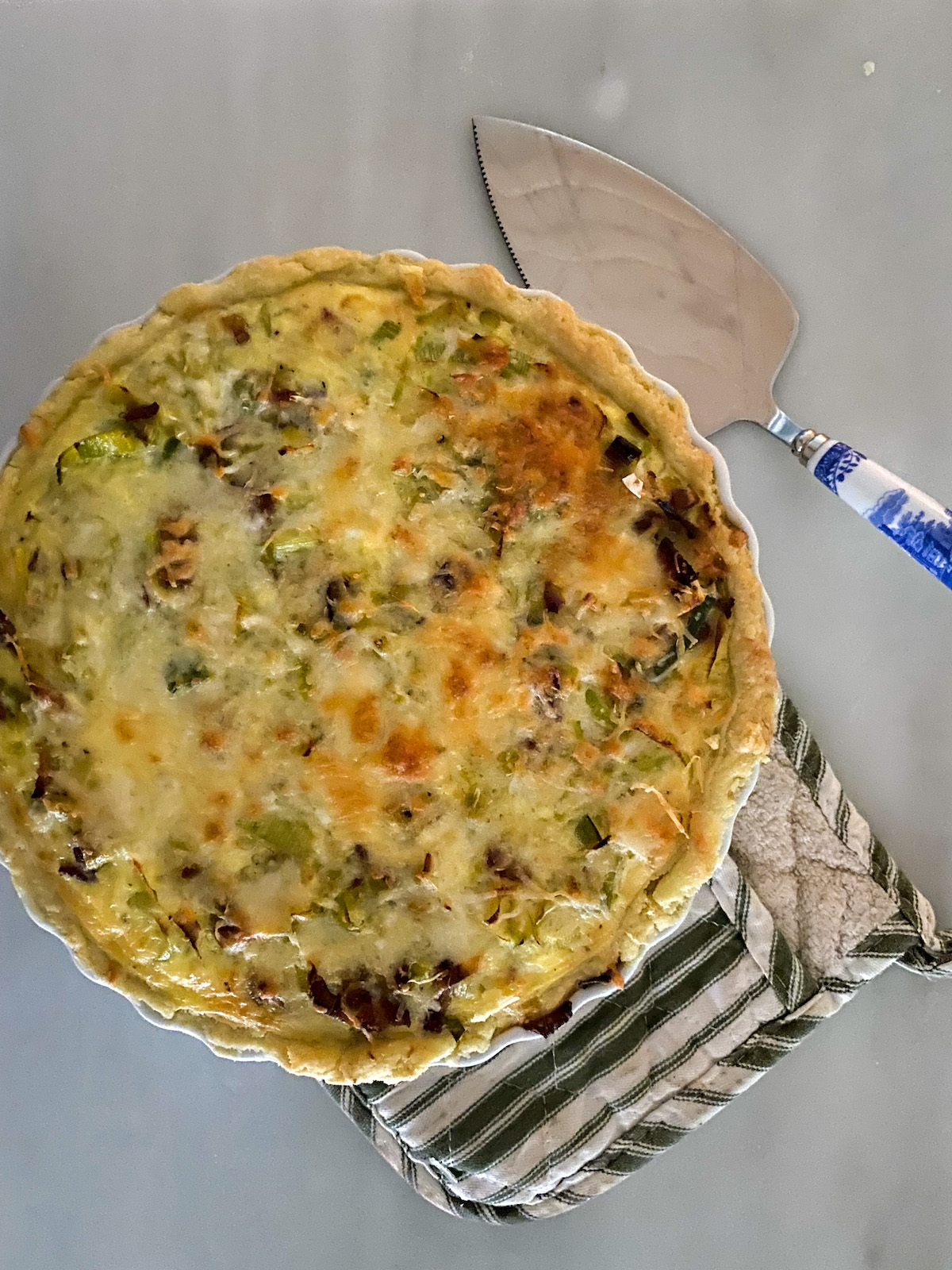 yellow green and creamy white birds eye view of quiche in a white ceramic dish on top of a green and white stripped pot holder adjacent to a blue and white handles serving utensil with a stainless steel triangular cake server white marble background