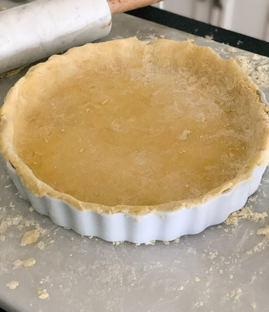 yellow pie crust in white ceramic dish white rolling pin with wooden handles on top of a white pastry board covered in flour white kitchen cabinets in the background 