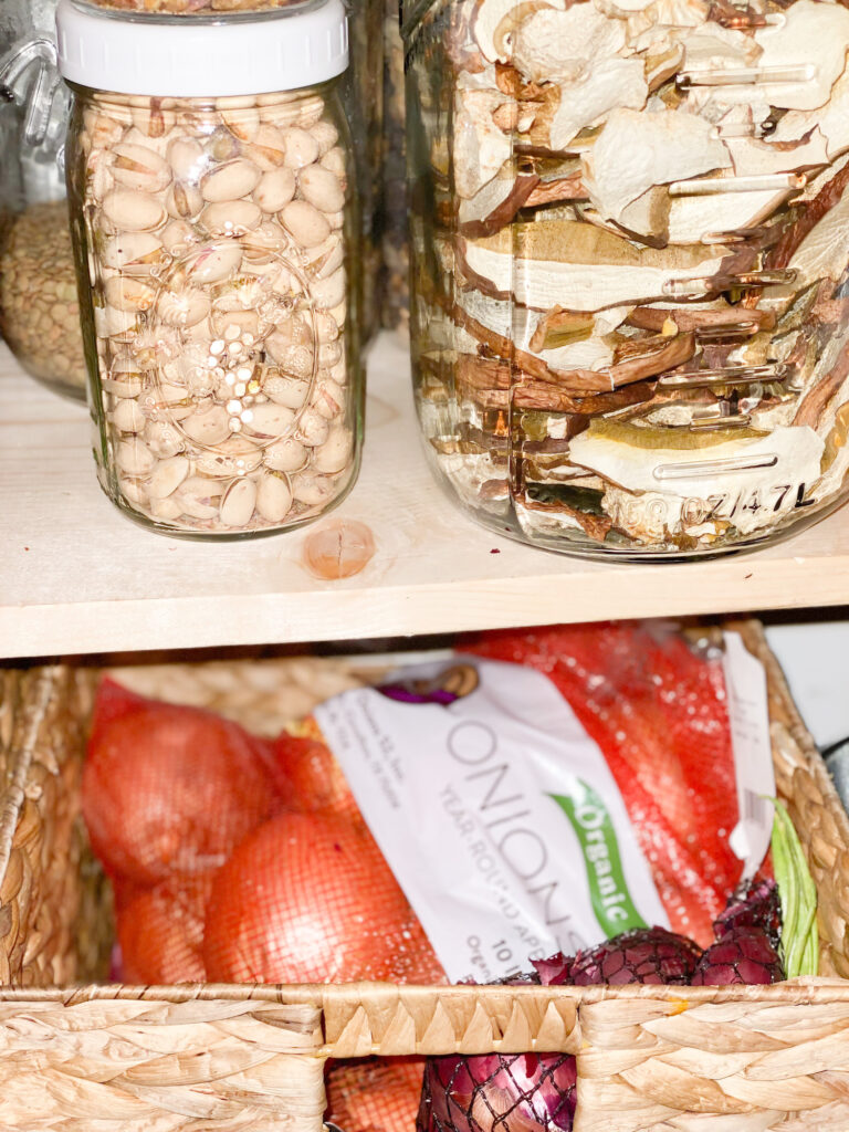 glass jar of pistachios in front of a glass jar filled with lentils nest to a glass jar filled with dried mushrooms all sitting on a pine board shelf. Below is a basket full of yellow onions