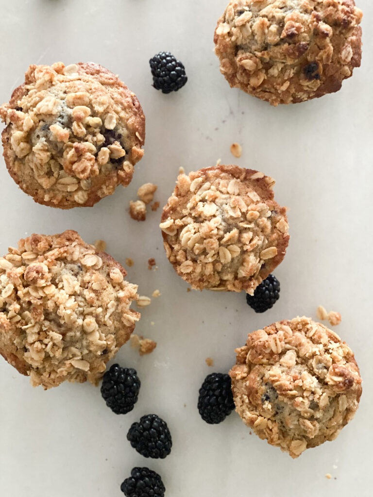 birds eye veweiw of five golden brown oatmeal muffins topped with oatmeal crumble, blackberries white background