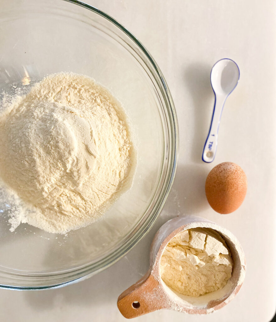 glass bowl full of Kamut® flour wooden measuring cup with more flour brown egg and blue and white teaspoon