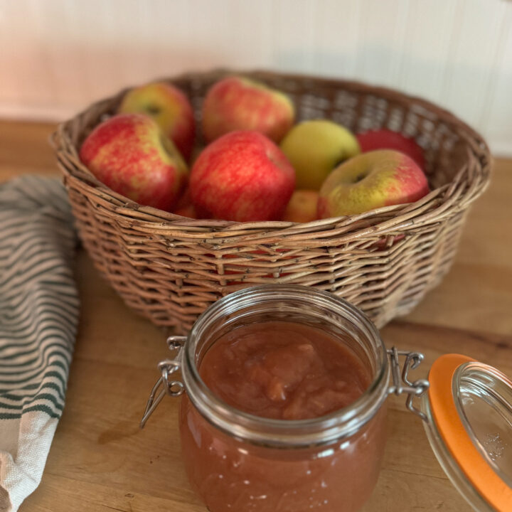 open glass jar of homemade apple sauce in front of a basket filled with apples to the left a green and white stripped tea towel